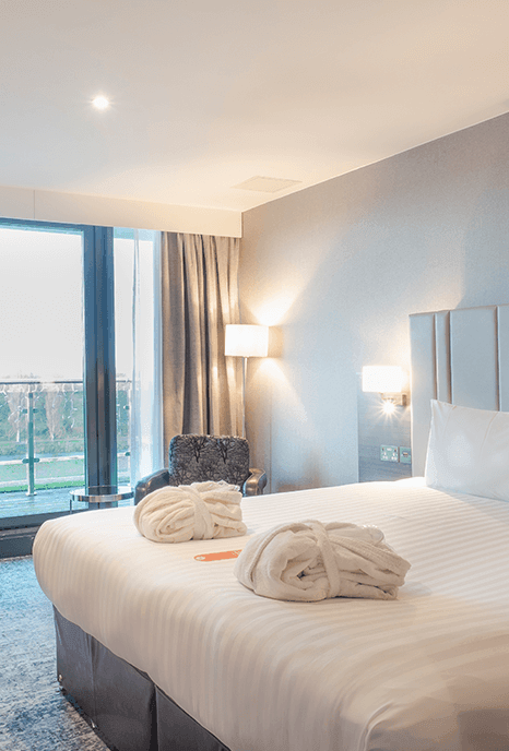 A modern hotel room with a large neatly-made bed, soft lighting, and a writing desk. Towels are folded on the bed, creating a welcoming ambiance.