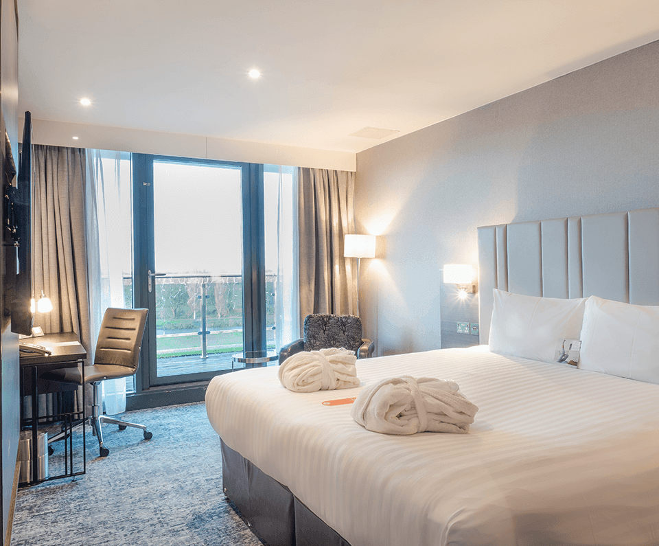 A modern hotel room with a large neatly-made bed, soft lighting, and a writing desk. Towels are folded on the bed, creating a welcoming ambiance.